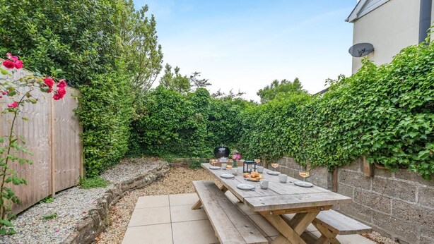 A patio with a wooden table set for dining, surrounded by a garden with a wooden fence and greenery. - 3 Bed in West Down (oc-r29627) (West Down)