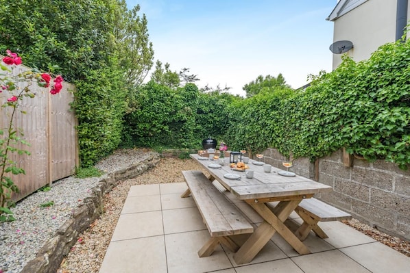 A patio with a wooden table set for dining, surrounded by a garden with a wooden fence and greenery.