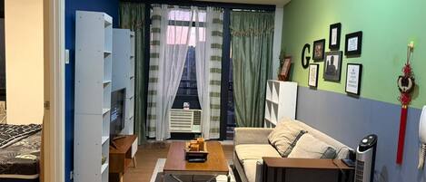 Una sala moderna con un sofá, una mesa de centro y un mueble para la televisión. Hay una cama en la habitación contigua, y una ventana con cortinas.