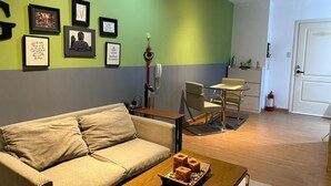 A living room with a sofa, coffee table, and a dining area with chairs and a table. There are framed pictures on the wall and a kitchen area in the background. - One Bedroom with Balcony City view  (Makati)