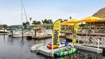 Un puerto deportivo con varias embarcaciones, una zona de alquiler de kayaks y un edificio con techo de paja.