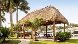 Ein Pavillon mit Strohdach und Picknicktischen, umgeben von Palmen und einem Yachthafen mit Booten.