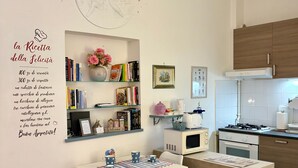 Una cucina moderna con zona pranzo, mobili bianchi, un forno a microonde, un fornello e una libreria con libri e oggetti decorativi.