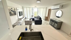 A modern living room with a sofa, a TV, a dining area, and a kitchen with a sink. - CB91-3 Bedrooms in beachfront complex (Carolina)