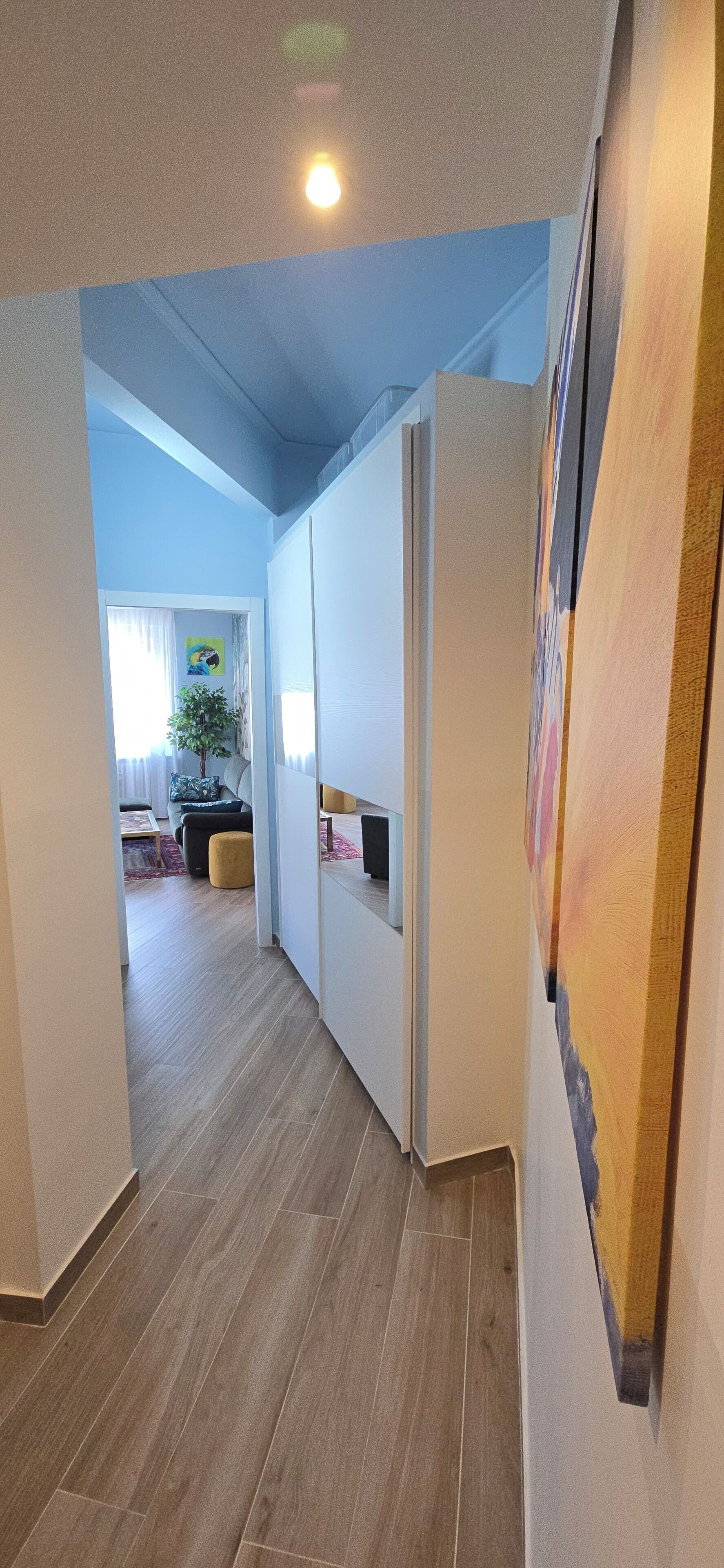Um corredor com chão de madeira, parede branca com um quadro e uma porta que dá para um quarto.