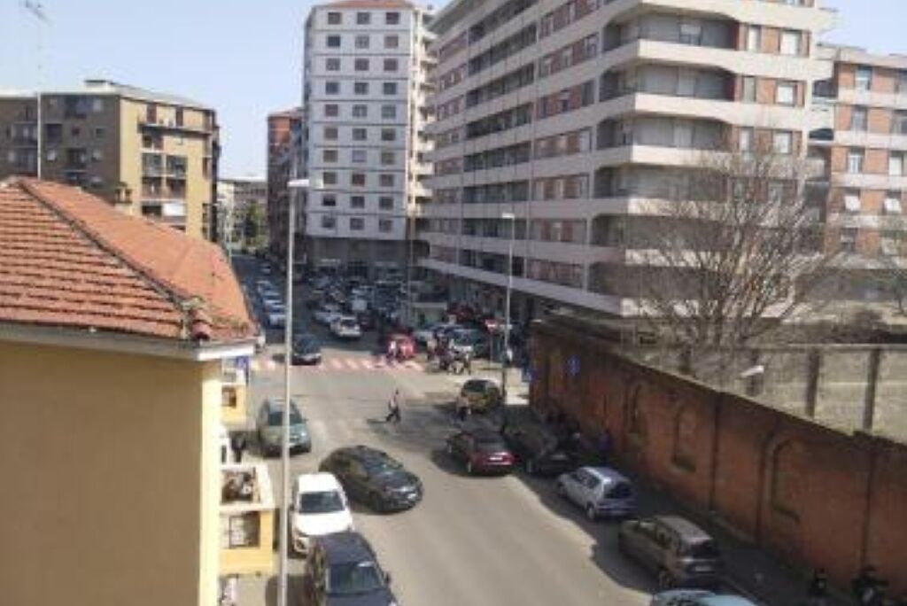 A multi-story building with a red-tiled roof, parked cars along the street, and a pedestrian crossing.