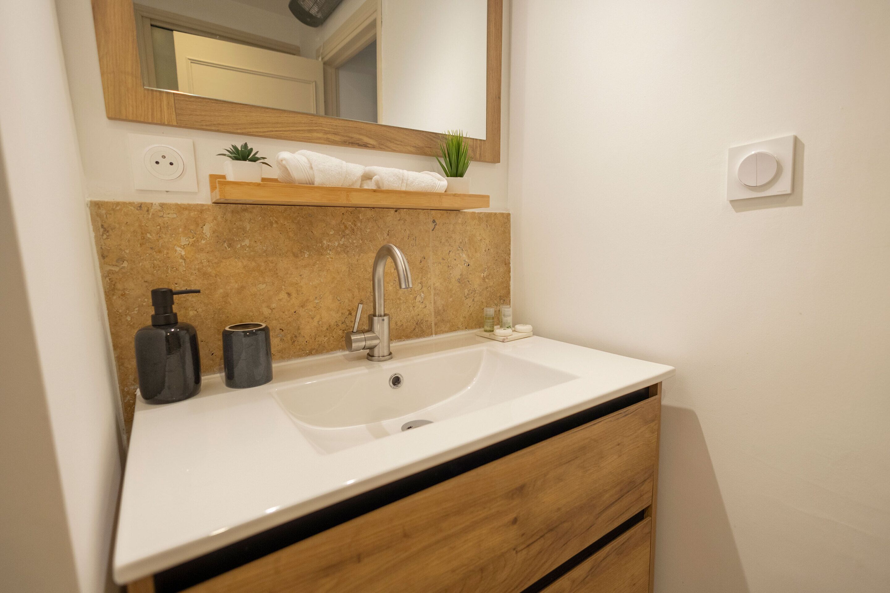 Un bagno con un lavandino bianco, un mobile bagno in legno, uno specchio, una piccola mensola con articoli da toilette e una pianta in vaso.