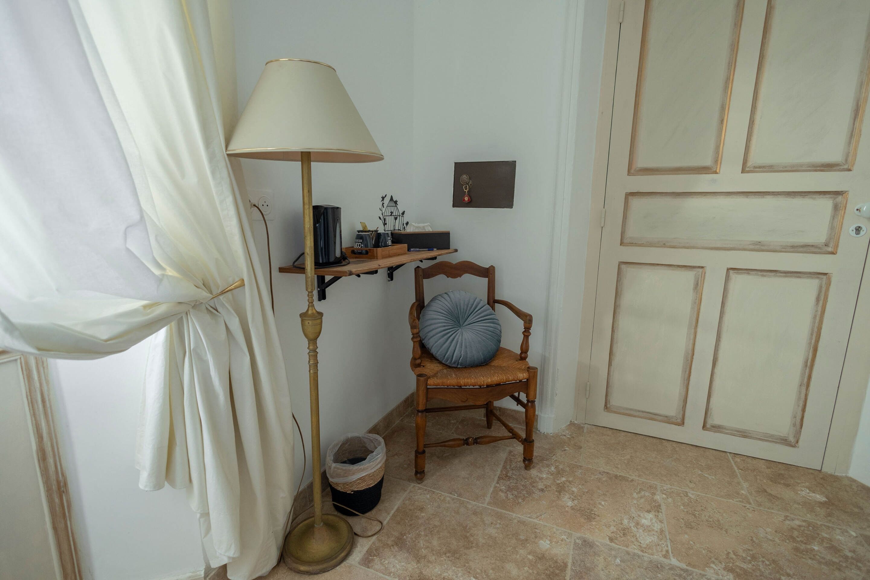 Una habitación con una silla de madera, una lámpara, una estantería con libros y una planta pequeña, además de una puerta con mirilla.