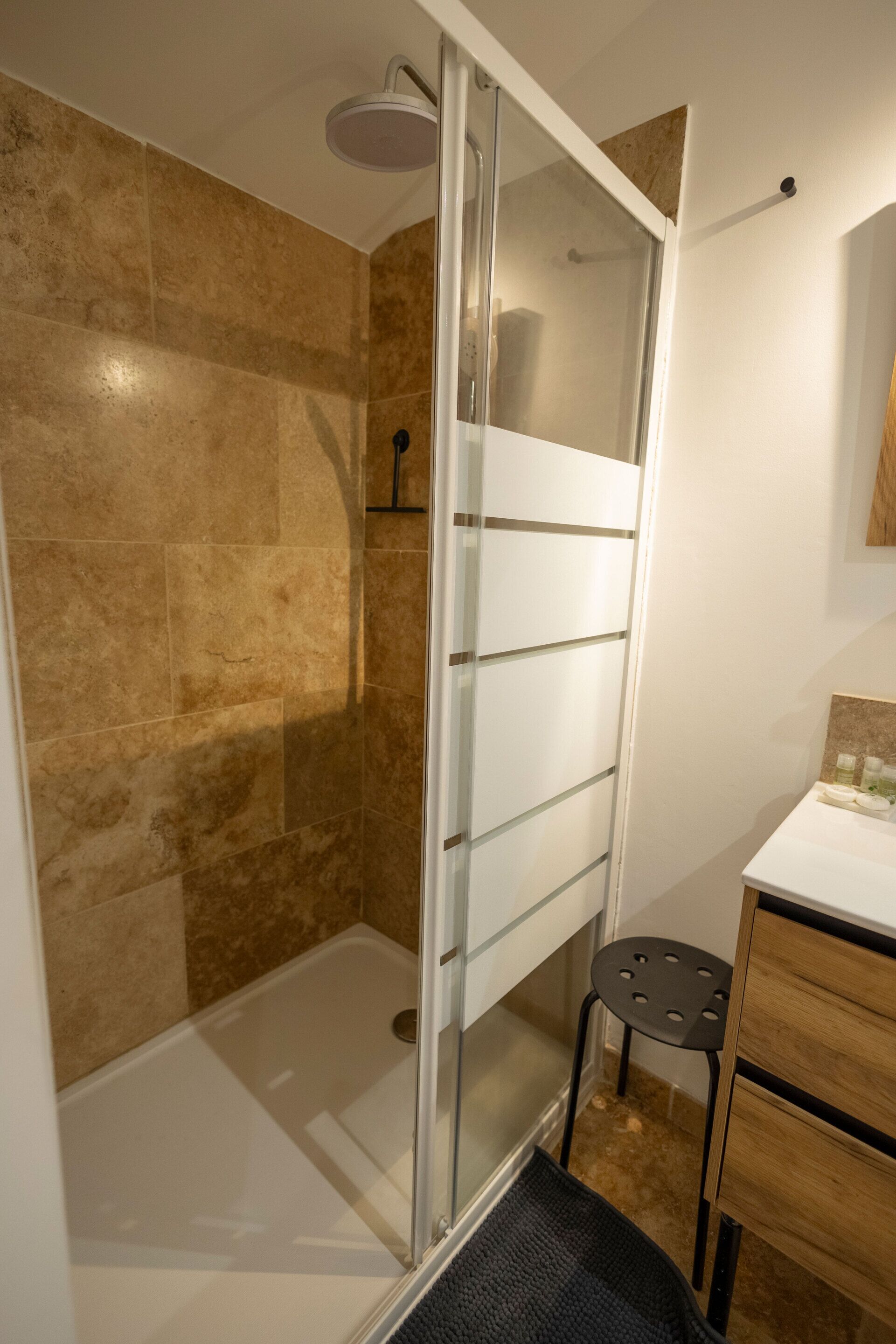 A modern bathroom with a glass shower enclosure, beige tiles, and a small wooden vanity.