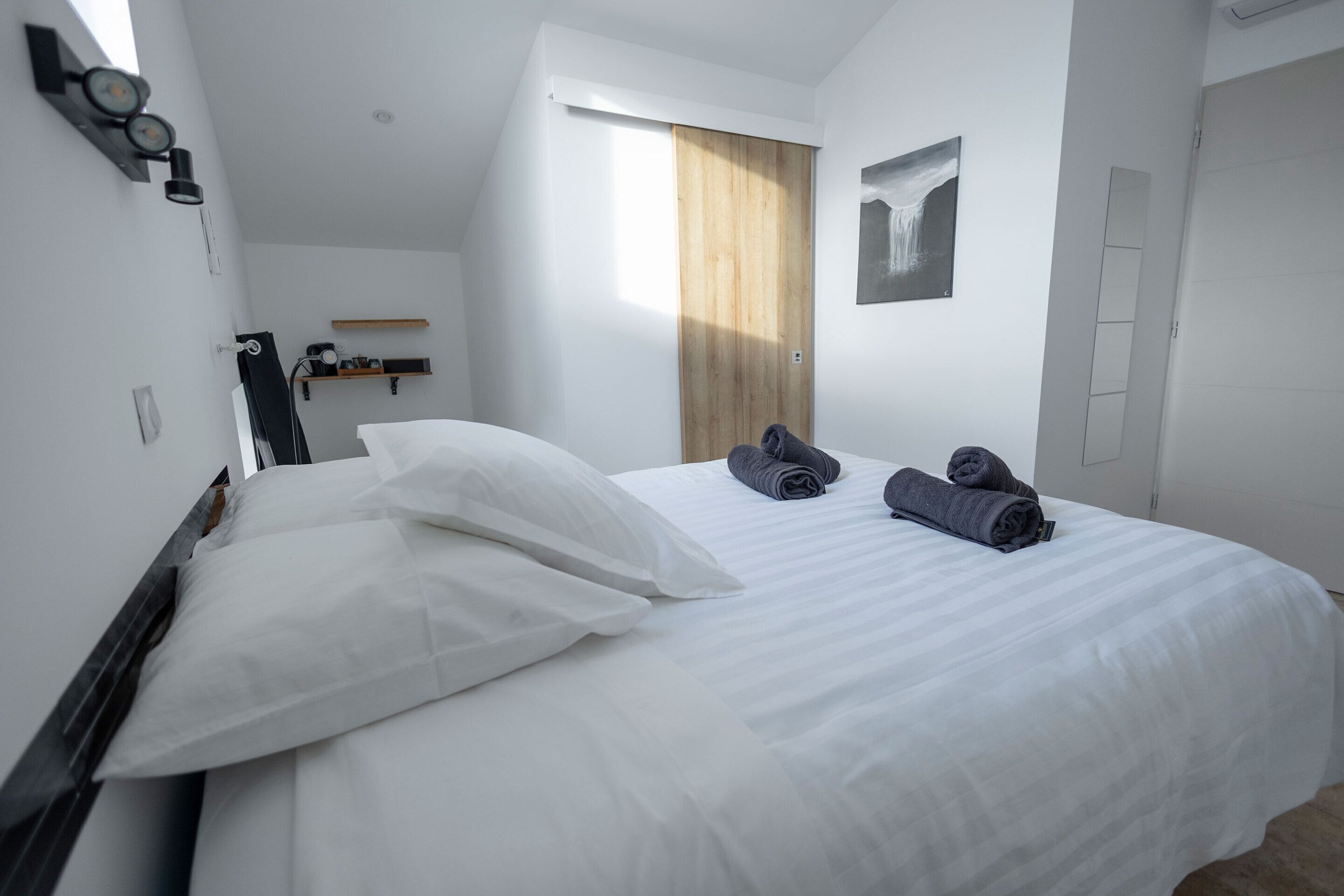 Une chambre d’hôtel avec un grand lit, du linge de lit blanc, une porte en bois, une fenêtre et une étagère fixée au mur.