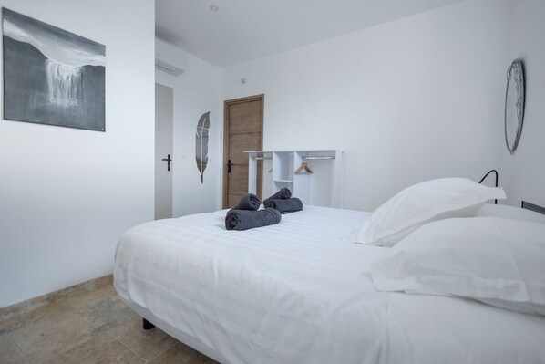 Una cama bien tendida con sábanas blancas y toallas grises, una puerta de madera y un perchero en la pared.