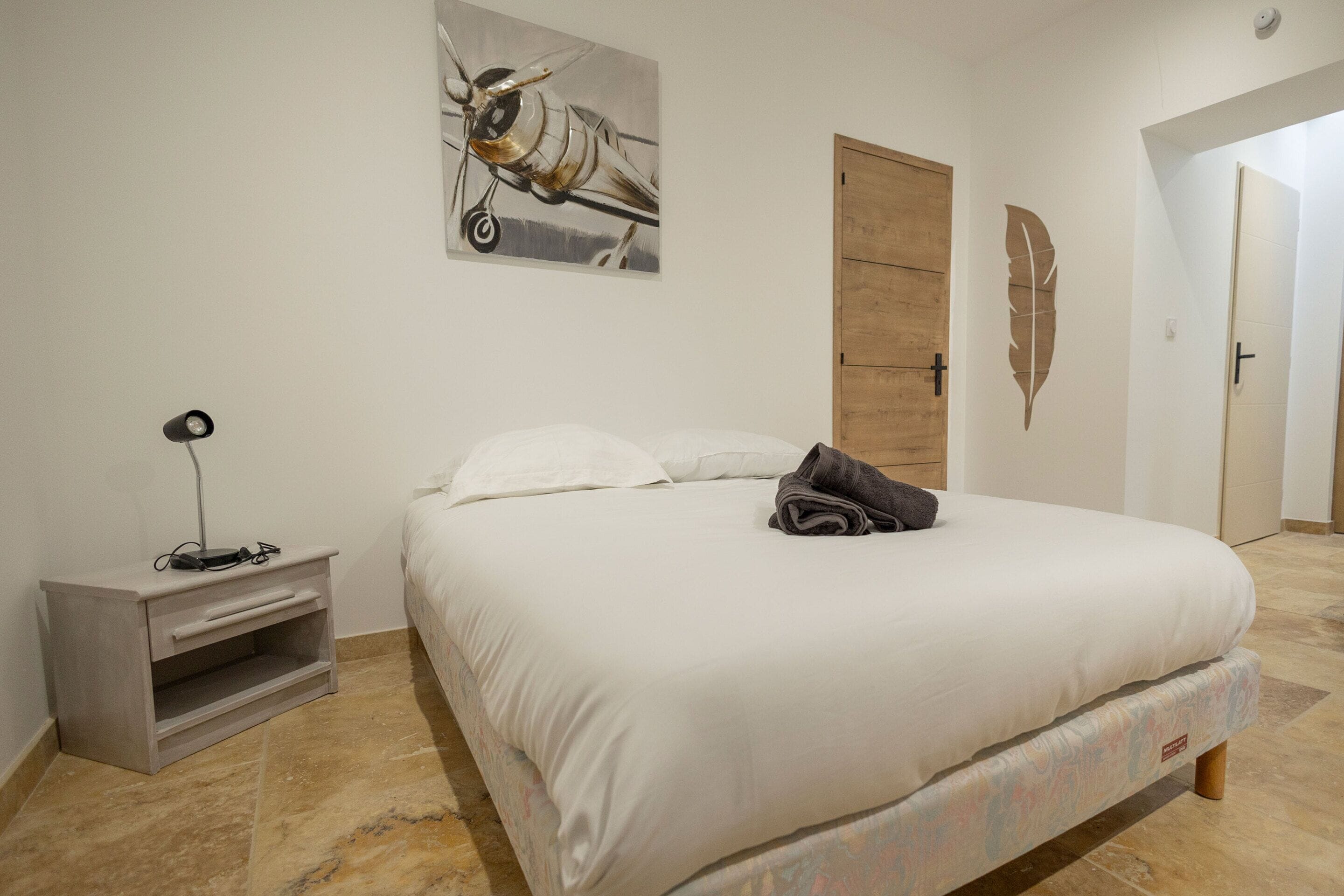 A neatly made bed with a pillow and a folded towel, a bedside table with a lamp, a wooden door, and a wall with a leaf decoration.