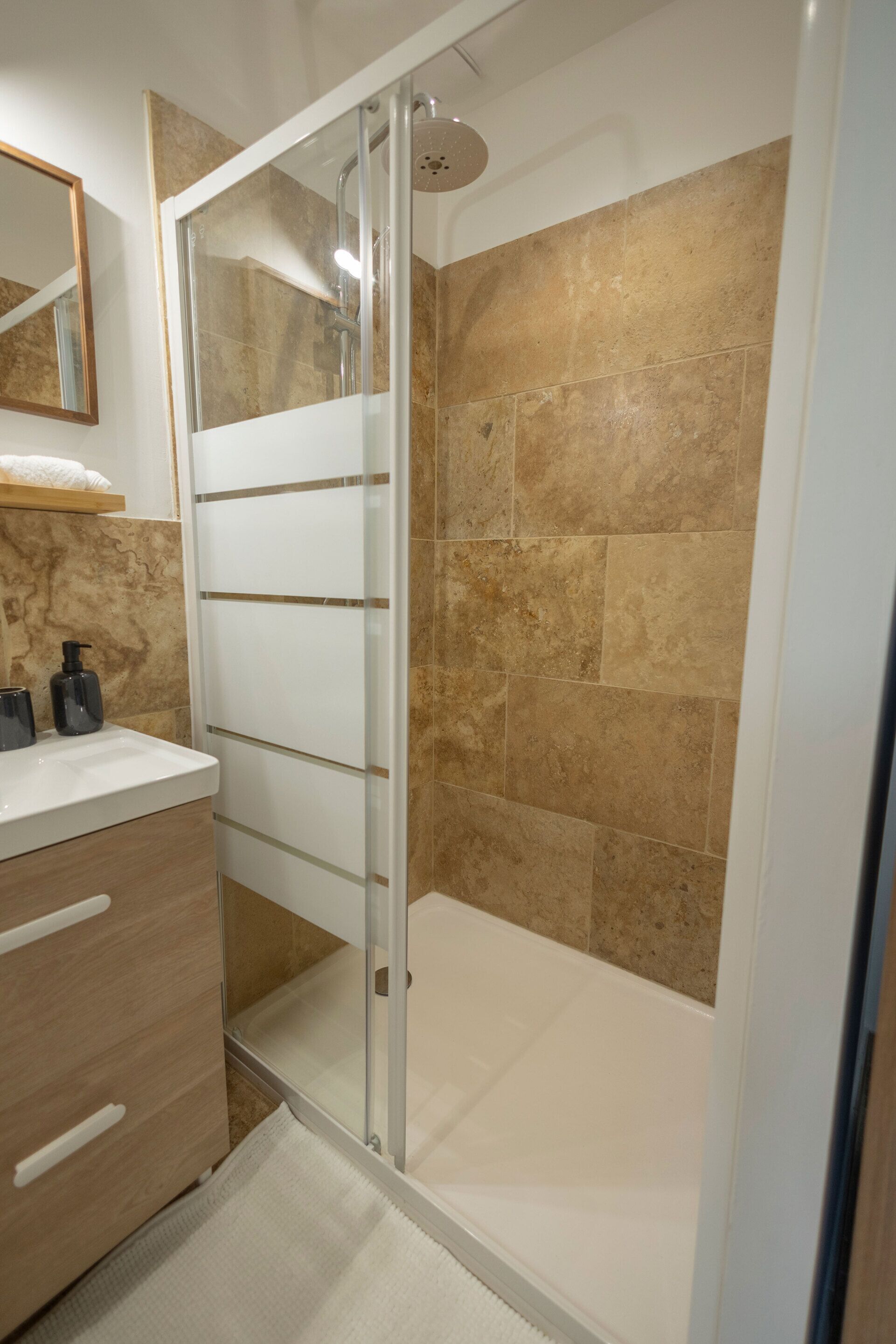 A modern bathroom with a glass-enclosed shower, beige tiles, a wooden vanity with drawers, and a mirror above the sink.