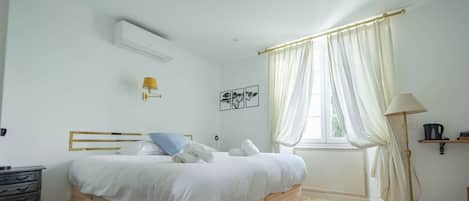 Un dormitorio con cama de madera, ropa de cama blanca, una mesita de noche, una lámpara, una ventana con cortinas y aire acondicionado instalado en la pared.