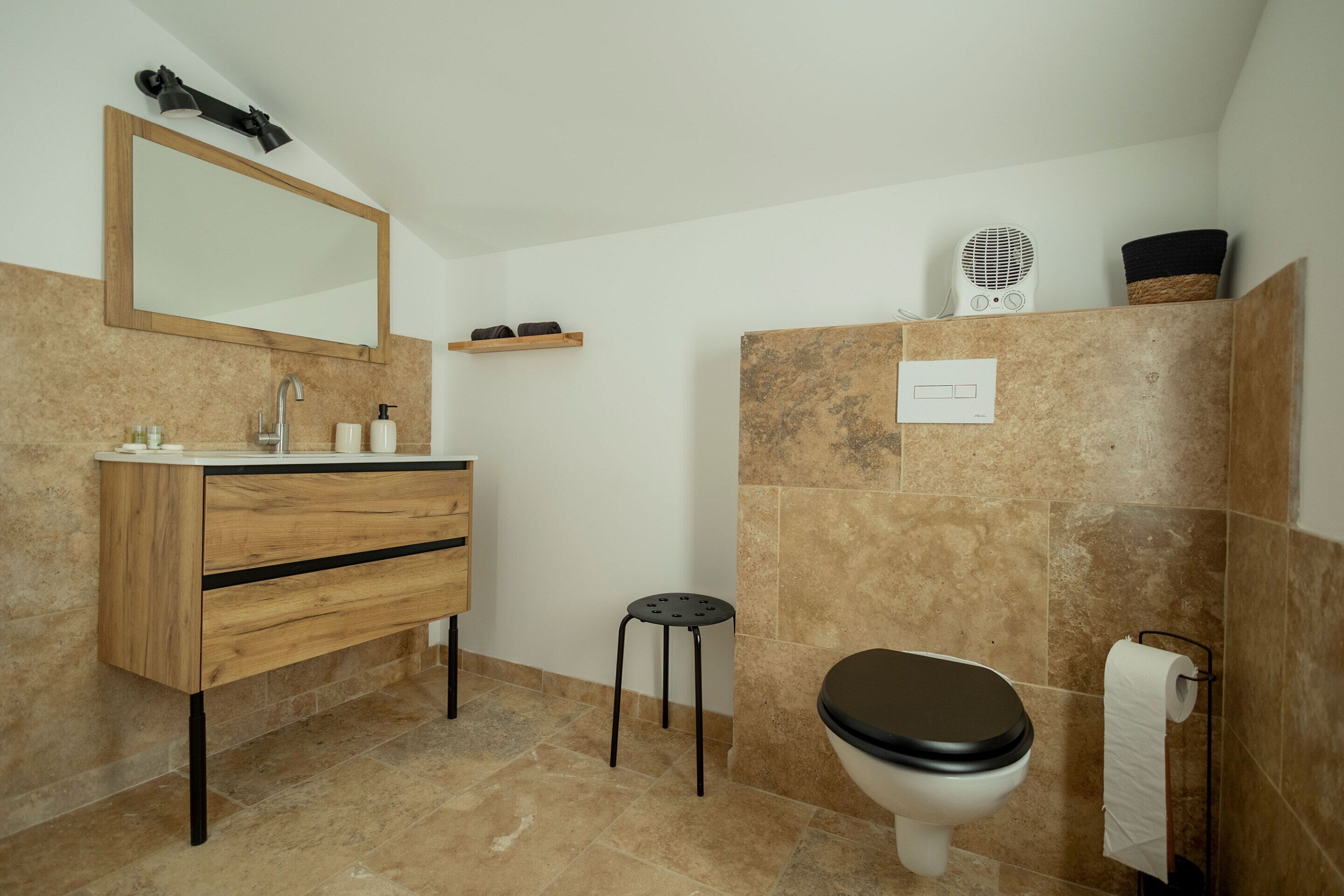 Uma casa de banho com toucador de madeira, sanita, um banquinho pequeno, espelho e secador de cabelo numa prateleira.