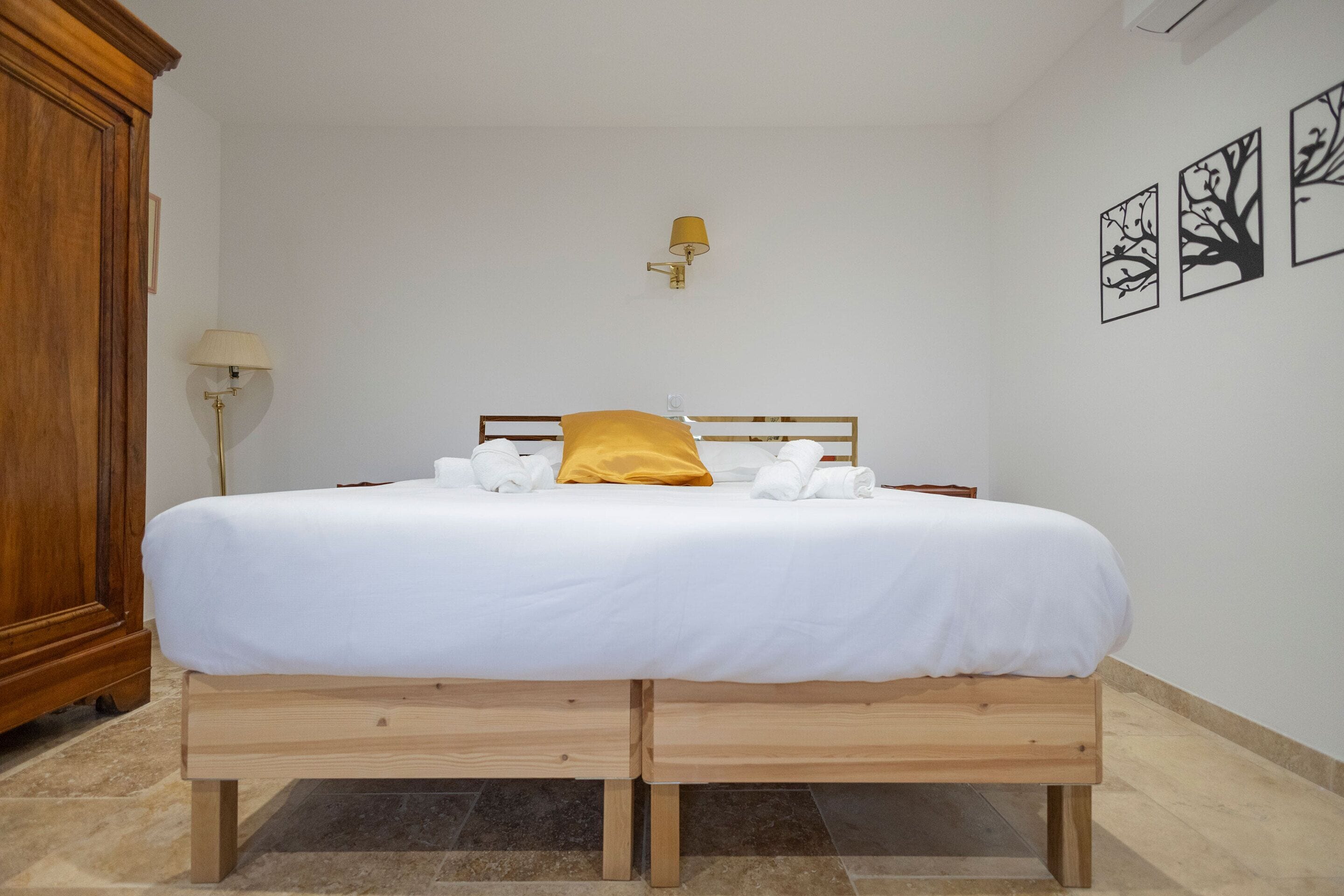 Una cama bien hecha con sábanas blancas y un cojín amarillo, una mesita de noche de madera y una lámpara de pared.