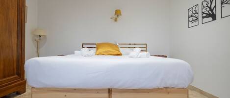 Una cama bien hecha con sábanas blancas y un cojín amarillo, una mesita de noche de madera y una lámpara de pared.