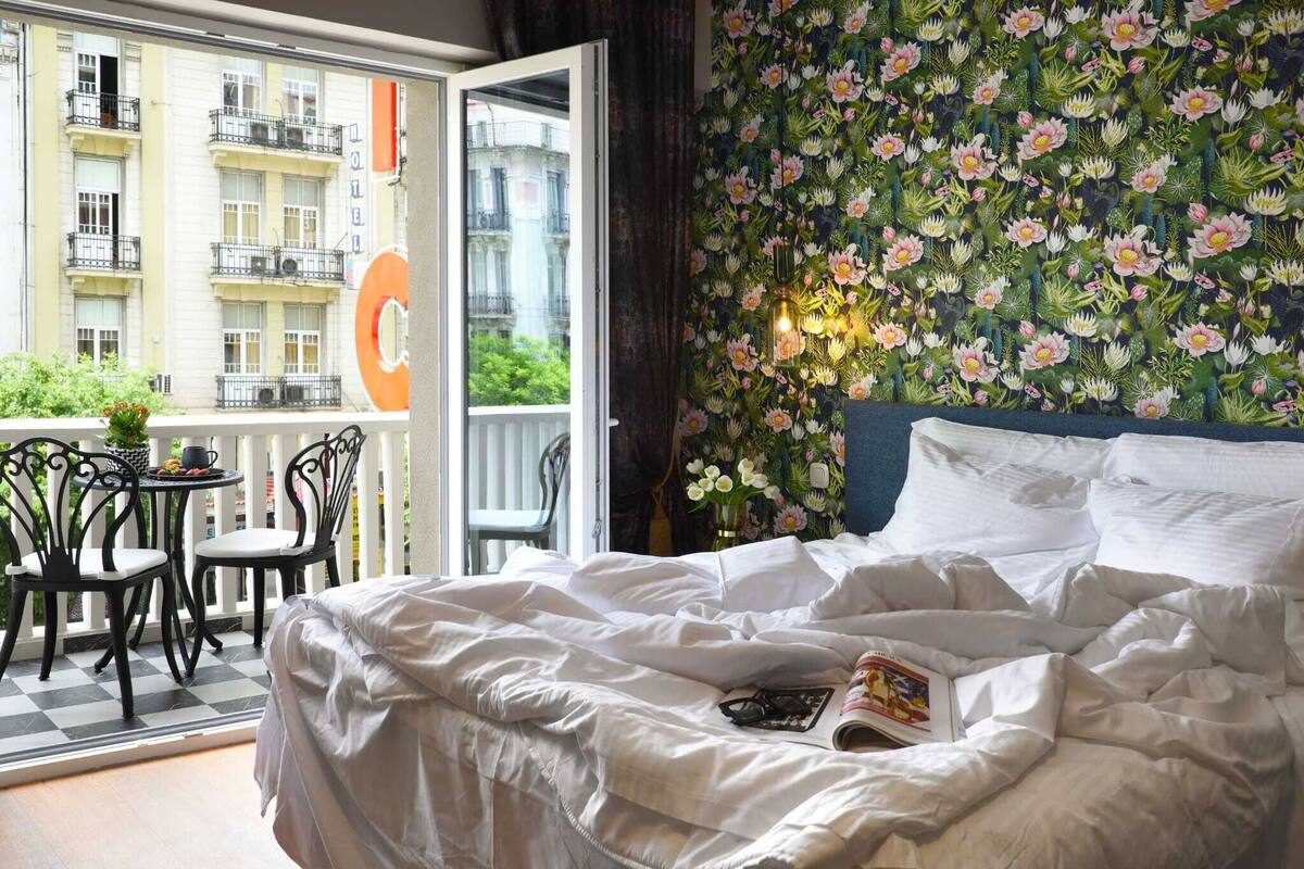 A hotel room with a bed, a balcony with a table and chairs, and a floral wallpaper.