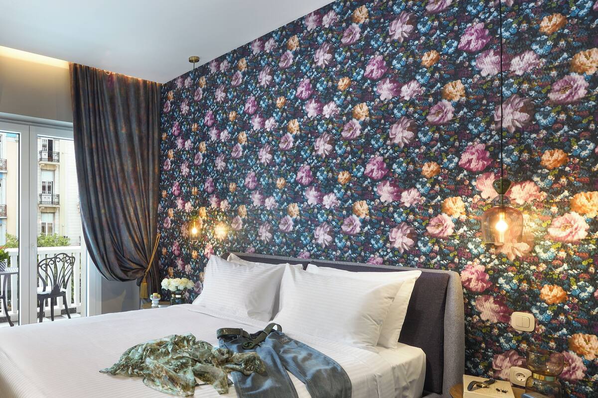 A bedroom with a floral wallpaper, a bed with white linens, a bedside table with a lamp, and a view of the outside balcony.