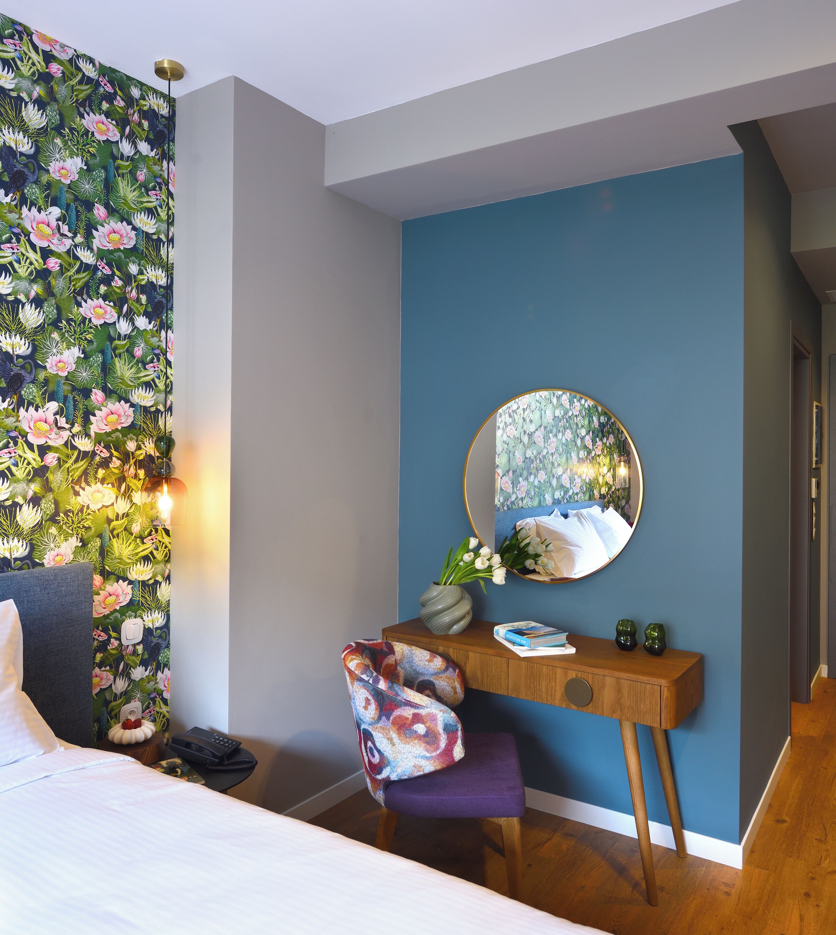 Une chambre avec un papier peint à motifs floraux, un miroir rond, un bureau en bois, une chaise violette et un lit.