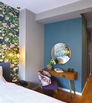 Quarto com papel de parede floral, um espelho redondo, escrivaninha de madeira, cadeira roxa e uma cama.