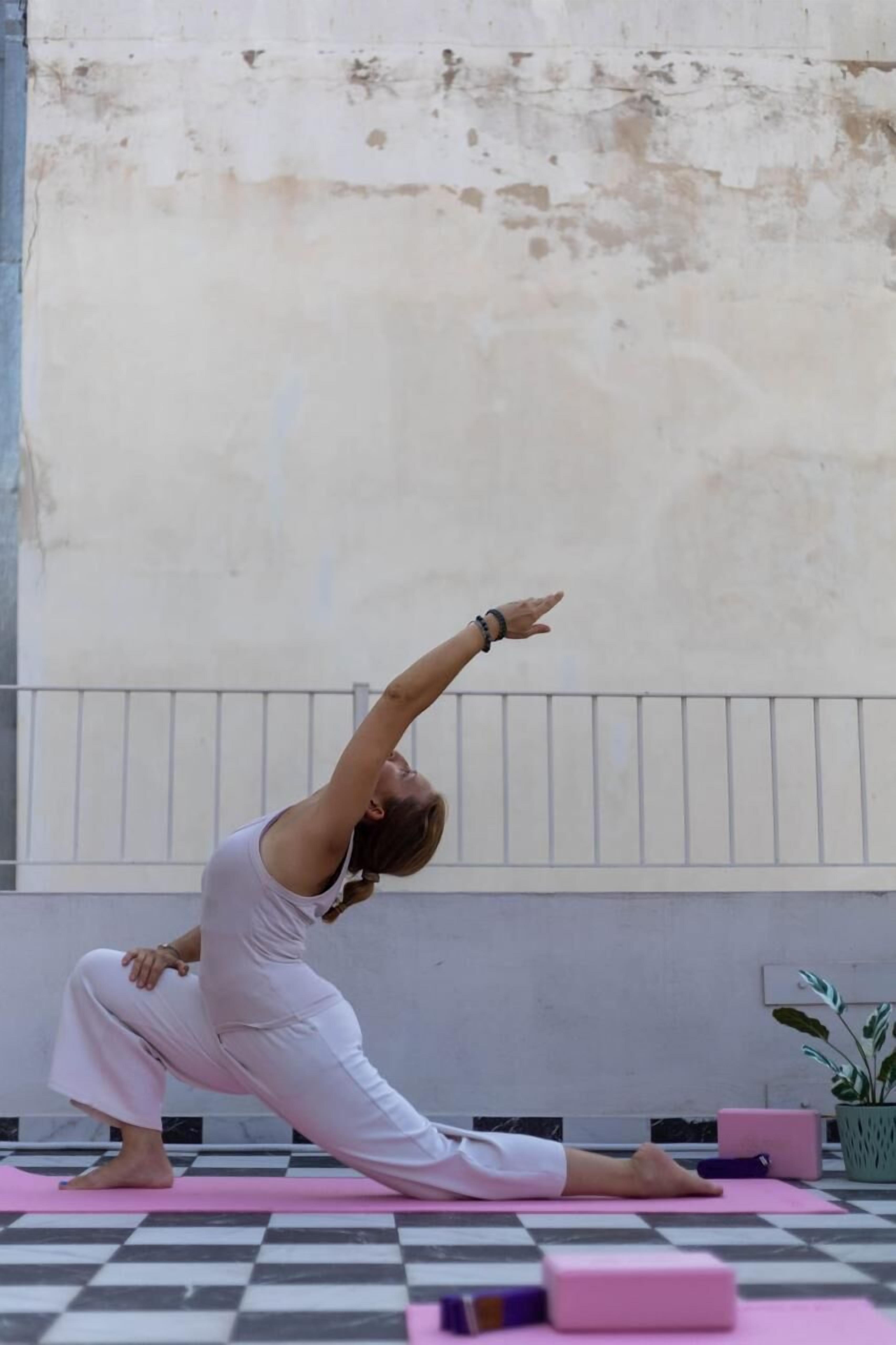 A person performing a yoga pose on a pink mat in an outdoor setting with a checkered floor and a plain wall in the background.