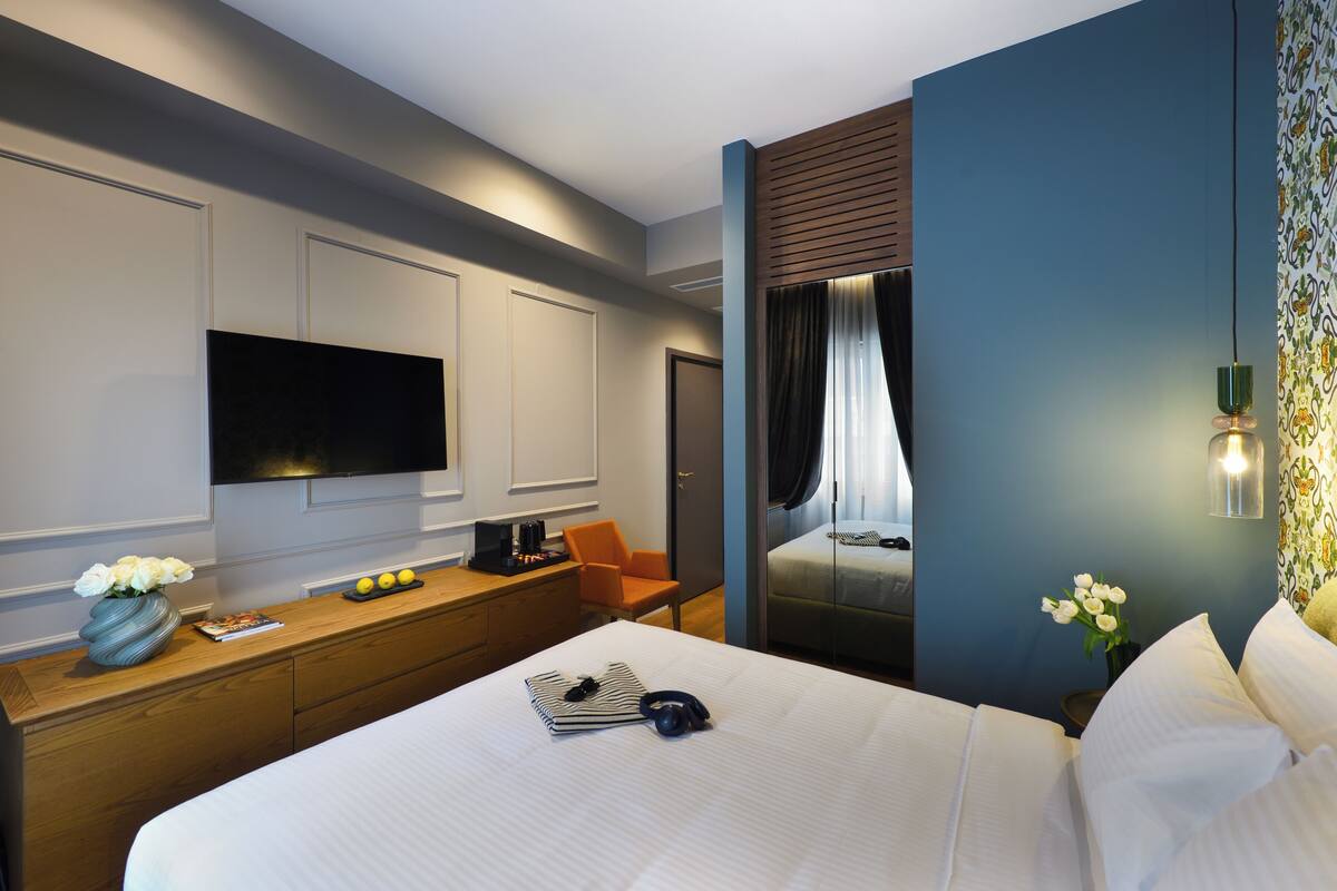 A modern hotel room with a large bed, a flat-screen TV, a wooden desk, and a view of a balcony.