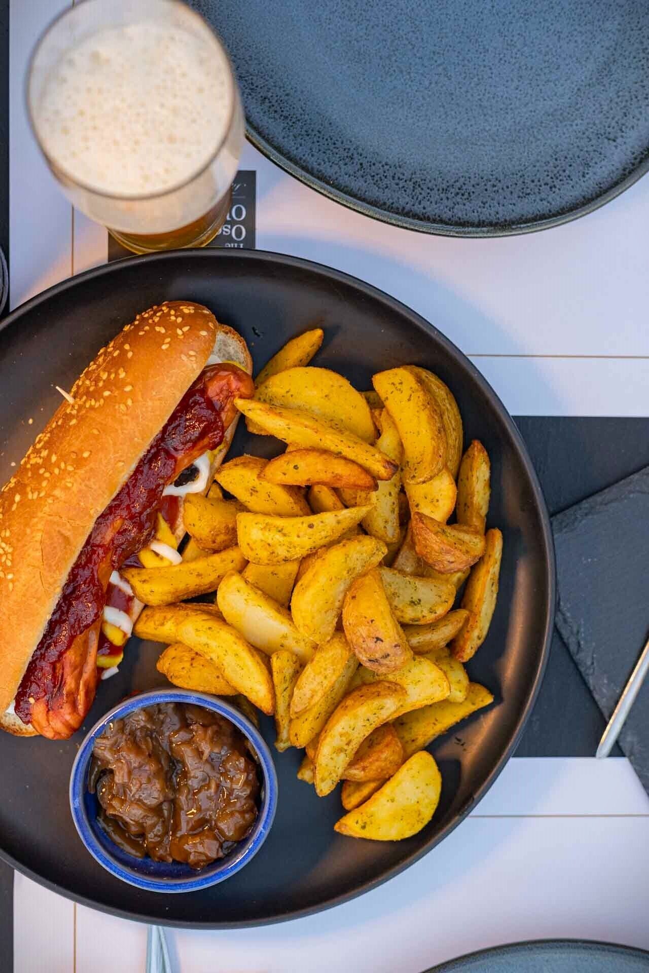 Lautasella on hot dog -sämpylä, ranskalaisia ja pieni kulho kastiketta.