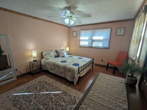 A bedroom with a bed, bedside lamps, a ceiling fan, a window with blinds, a chair, and a potted plant.