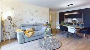Een moderne woonkamer met een grijze bank, een ronde glazen salontafel, en een eethoek met een donkere keuken op de achtergrond.