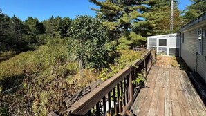 Une terrasse en bois mène à une maison mobile entourée d’une clôture en métal, elle-même bordée d’une forêt dense et d’un grand arbre à feuilles persistantes.