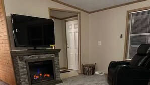 Un salon doté d’une cheminée en pierre, d’un téléviseur à écran plat fixé au-dessus, d’un fauteuil inclinable en cuir et d’une porte donnant sur une autre pièce.