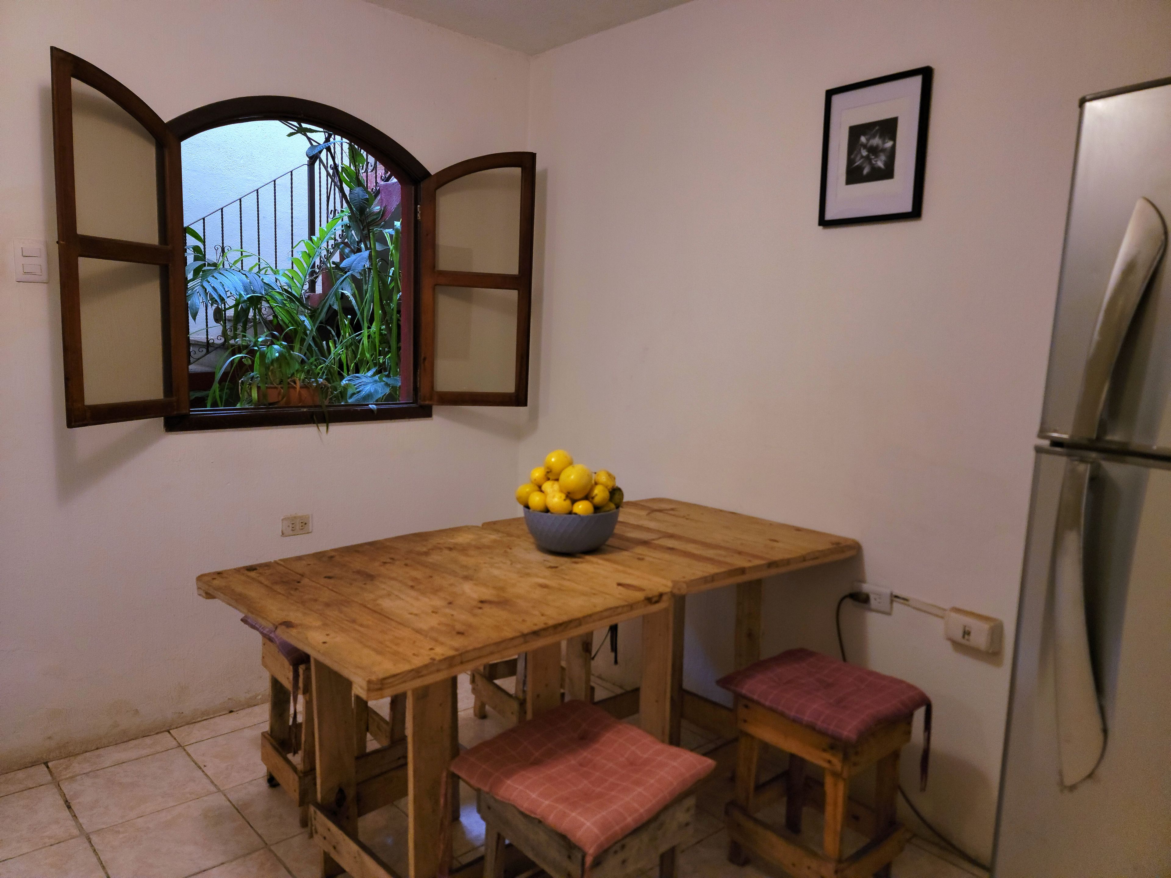 A kitchen with a wooden table, two stools, a bowl of lemons, a window with plants, and a refrigerator.