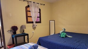 A room with two beds, a small table with a lamp, a window with a curtain, and a wall-mounted mirror.