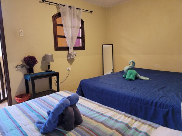 A room with two beds, a small table with a lamp, a window with a curtain, and a wall-mounted mirror. - Castillo De Los Gigantes (Antigua Guatemala)