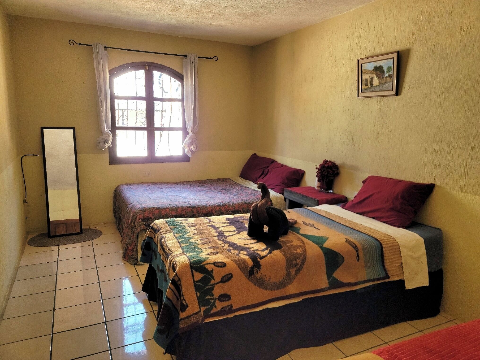 Quarto com duas camas, janela com cortinas, quadro na parede e espelho de corpo inteiro.