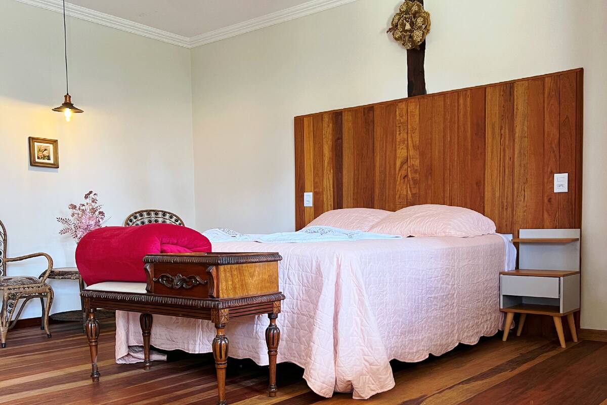 A bedroom with a wooden headboard, a bed with pink bedding, a wooden nightstand, a chair, and a hanging light fixture.