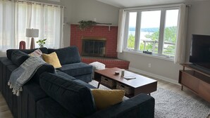 A living room with a dark-colored sofa, a fireplace, a television, and large windows offering a view of the outdoors.
