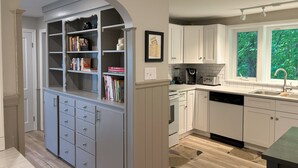A kitchen with white cabinets, a built-in bookshelf, a window with a view of trees, and a countertop with a coffee maker.