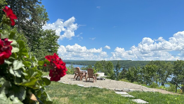 A gravel patio with Adirondack chairs overlooking a lake, surrounded by greenery and trees.