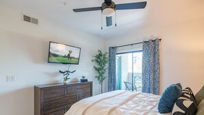 A bedroom with a bed, a dresser, a television on the wall, a ceiling fan, and a window with curtains.
