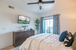 A bedroom with a bed, a dresser, a television on the wall, a ceiling fan, and a window with curtains.