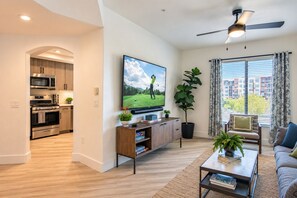 A modern living room with a flat-screen TV, a ceiling fan, a large window with curtains, a sofa, and a coffee table. - Heated Pool | Hot Tub | Walk to Kierland | NearTPC (Scottsdale)
