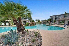 A pool area with palm trees, lounge chairs, and a building with balconies.