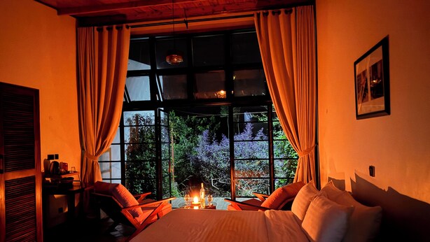 A cozy bedroom with a large bed, a table with candles, and a view of greenery outside. - IGWARA LODGE (Karuma)