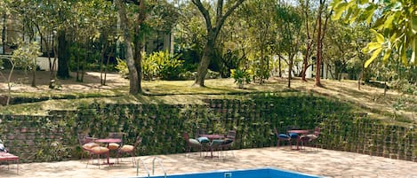 En swimmingpool med fliseterrasse, omgivet af en have med forskellige planter og træer.