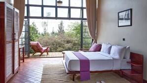 A bedroom with a bed, a chair, and a view of greenery through large windows. - IGWARA LODGE (Karuma)