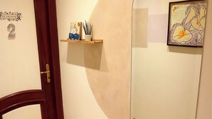 A hallway with a wooden door labeled "Bedroom," a small shelf with decorative items, and a large mirror reflecting the room.