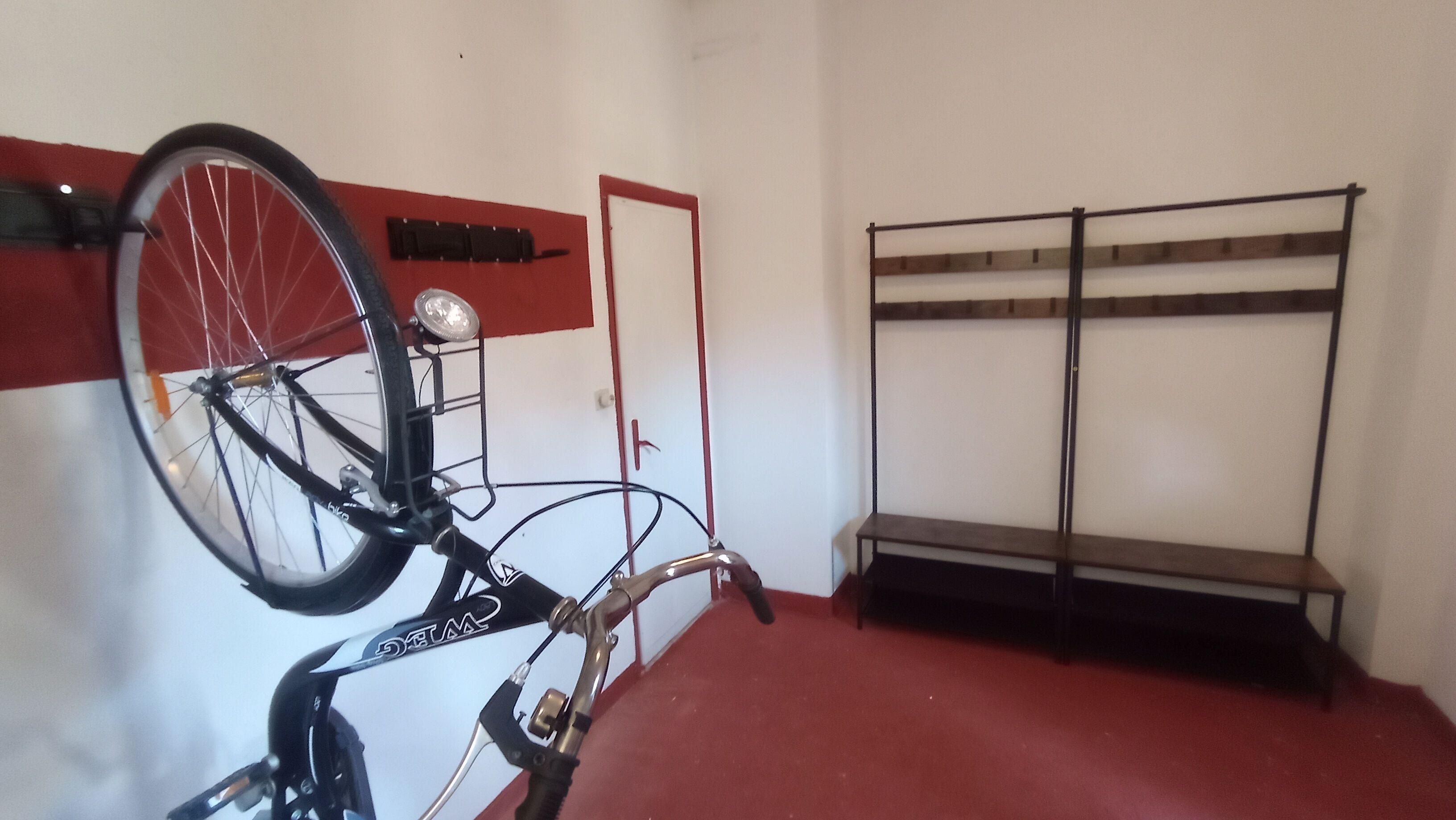 A bicycle with a front basket and a rear rack, a red-framed door, and a metal bench against a white wall.