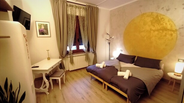 A bedroom with a large bed, a desk, a chair, a bedside table, and a window with curtains.